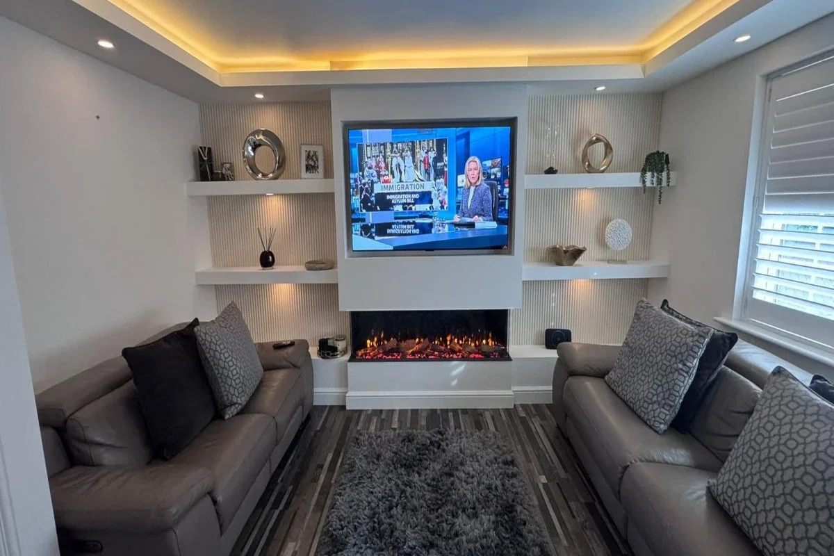 The image shows a modern living room centred around a bespoke media wall Sheffield installation. The media wall forms a striking focal point, combining clean white cabinetry with warm lighting and textured finishes. At the centre of the wall is a large flat-screen television, neatly inset within a white surround. Below the TV sits a sleek, built-in electric fireplace with glowing ember effects, adding warmth and ambience. Surrounding the central section are symmetrical floating shelves on both sides, used to display decorative items such as framed photos, sculptural ornaments, small plants, and vases. The back panels behind the shelving feature vertical fluted wall detailing, enhancing texture and depth. Warm LED lighting is integrated into the shelves and ceiling recess, creating a soft, inviting glow throughout the space. In the foreground, two contemporary sofas face one another, upholstered in neutral tones, complemented by patterned cushions. A grey shag rug sits centrally on a wood-effect floor, tying the room together. To the right, large windows fitted with plantation shutters allow natural light to balance the artificial lighting. Overall, the design is minimalist yet luxurious, with a cohesive palette of whites, greys, and warm lighting accents.