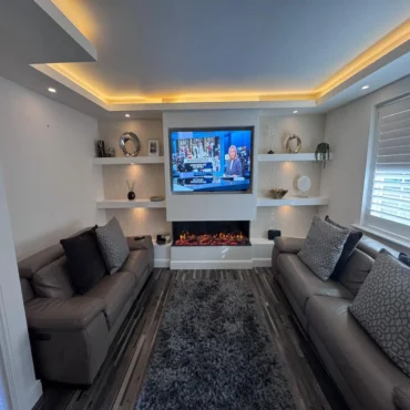 The image shows a modern living room centred around a bespoke media wall Sheffield installation. The media wall forms a striking focal point, combining clean white cabinetry with warm lighting and textured finishes. At the centre of the wall is a large flat-screen television, neatly inset within a white surround. Below the TV sits a sleek, built-in electric fireplace with glowing ember effects, adding warmth and ambience. Surrounding the central section are symmetrical floating shelves on both sides, used to display decorative items such as framed photos, sculptural ornaments, small plants, and vases. The back panels behind the shelving feature vertical fluted wall detailing, enhancing texture and depth. Warm LED lighting is integrated into the shelves and ceiling recess, creating a soft, inviting glow throughout the space. In the foreground, two contemporary sofas face one another, upholstered in neutral tones, complemented by patterned cushions. A grey shag rug sits centrally on a wood-effect floor, tying the room together. To the right, large windows fitted with plantation shutters allow natural light to balance the artificial lighting. Overall, the design is minimalist yet luxurious, with a cohesive palette of whites, greys, and warm lighting accents.