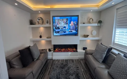 The image shows a modern living room centred around a bespoke media wall Sheffield installation. The media wall forms a striking focal point, combining clean white cabinetry with warm lighting and textured finishes. At the centre of the wall is a large flat-screen television, neatly inset within a white surround. Below the TV sits a sleek, built-in electric fireplace with glowing ember effects, adding warmth and ambience. Surrounding the central section are symmetrical floating shelves on both sides, used to display decorative items such as framed photos, sculptural ornaments, small plants, and vases. The back panels behind the shelving feature vertical fluted wall detailing, enhancing texture and depth. Warm LED lighting is integrated into the shelves and ceiling recess, creating a soft, inviting glow throughout the space. In the foreground, two contemporary sofas face one another, upholstered in neutral tones, complemented by patterned cushions. A grey shag rug sits centrally on a wood-effect floor, tying the room together. To the right, large windows fitted with plantation shutters allow natural light to balance the artificial lighting. Overall, the design is minimalist yet luxurious, with a cohesive palette of whites, greys, and warm lighting accents.