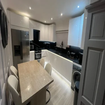 Modern white kitchen with black countertops, stainless steel fridge, built-in oven, washing machine, under-cabinet lighting, and a small dining table with four chairs.