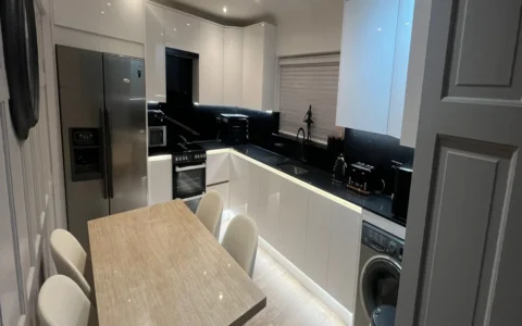 Modern white kitchen with black countertops, stainless steel fridge, built-in oven, washing machine, under-cabinet lighting, and a small dining table with four chairs.