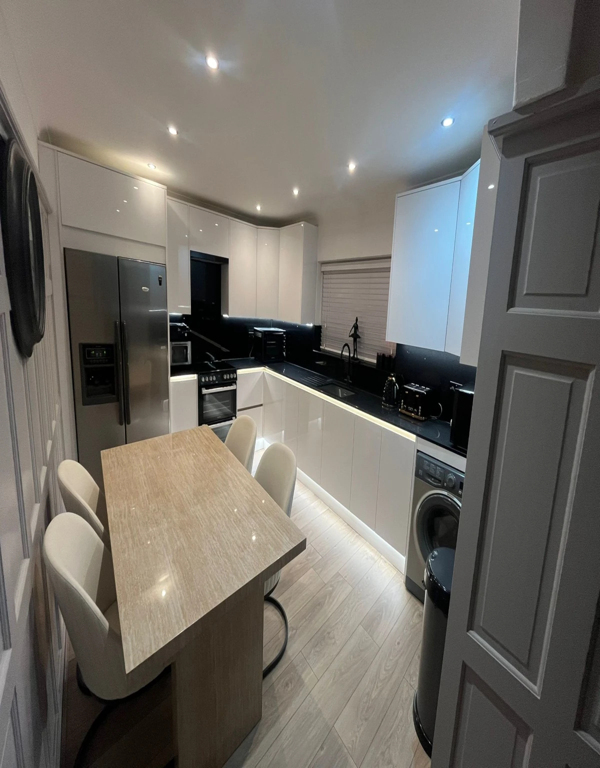 Modern white kitchen with black countertops, stainless steel fridge, built-in oven, washing machine, under-cabinet lighting, and a small dining table with four chairs.